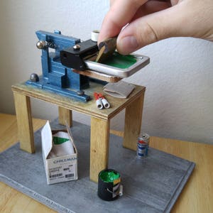 May include: A miniature screen printing press on a wooden table with a green screen, a box of Chillman brand supplies, a can of paint, and a roll of tape.