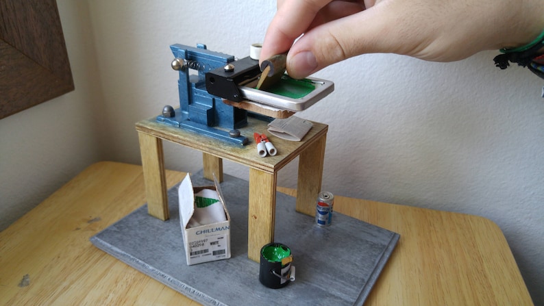 May include: A miniature screen printing press on a wooden table with a green screen, a box of Chillman brand supplies, a can of paint, and a roll of tape.