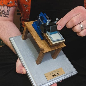 May include: A miniature wooden table with a blue and black printing press on top. The table is on a gray and silver plaque with a gold plaque that reads "Board of Directors, Student Member 2019-2020, BOD by Galaxy".