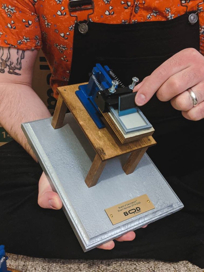 May include: A miniature wooden table with a blue and black printing press on top. The table is on a gray and silver plaque with a gold plaque that reads "Board of Directors, Student Member 2019-2020, BOD by Galaxy".