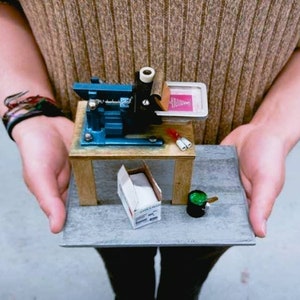 May include: A miniature screen printing press on a wooden table with a small box of supplies and a green paint can. The press is blue and has a pink screen with a design on it.