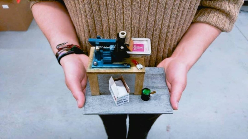 May include: A miniature screen printing press on a wooden table with a small box of supplies and a green paint can. The press is blue and has a pink screen with a design on it.