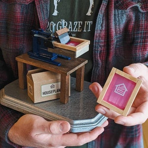 May include: A small wooden printing press with a blue metal frame and a brown cardboard box labeled "HOUSEPLAN". A pink screen printing frame with a white house icon is held in the right hand.