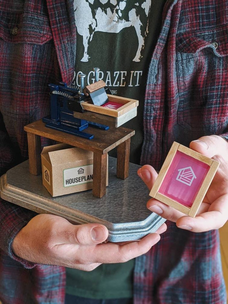 May include: A small wooden printing press with a blue metal frame and a brown cardboard box labeled "HOUSEPLAN". A pink screen printing frame with a white house icon is held in the right hand.