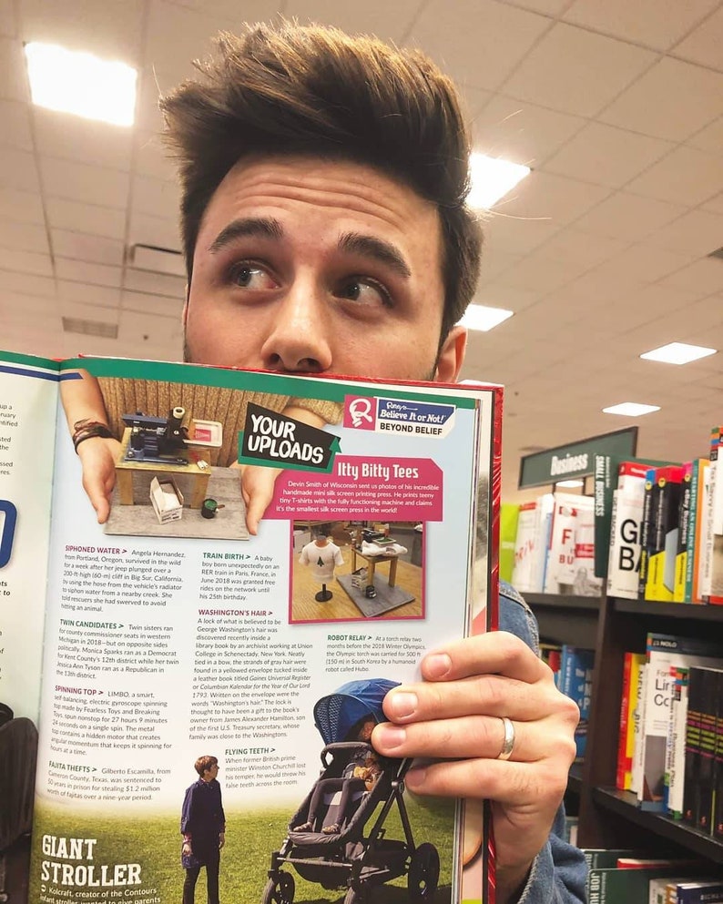May include: A man is holding open a magazine with a spread about unusual facts. The magazine features a photo of a tiny T-shirt printed on a tiny screen, a photo of a spinning top, and a photo of a robot relay race. The magazine is titled "Believe It or Not! Beyond Belief".