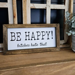 May include: A rectangular wooden framed sign with the words "BE HAPPY!" in bold black letters, followed by "bitches hate that" in a smaller font. The sign is set against a white background.