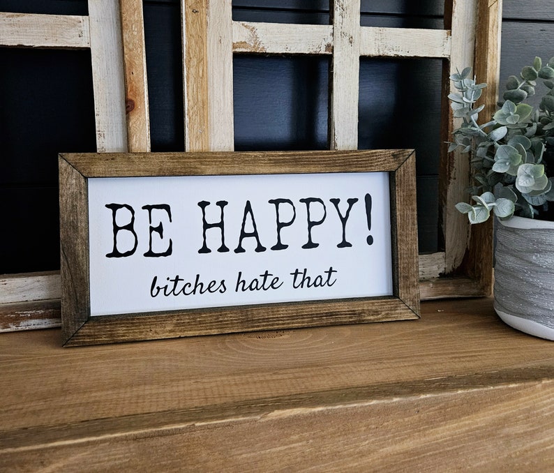 May include: A rectangular wooden framed sign with the words "BE HAPPY!" in bold black letters, followed by "bitches hate that" in a smaller font. The sign is set against a white background.