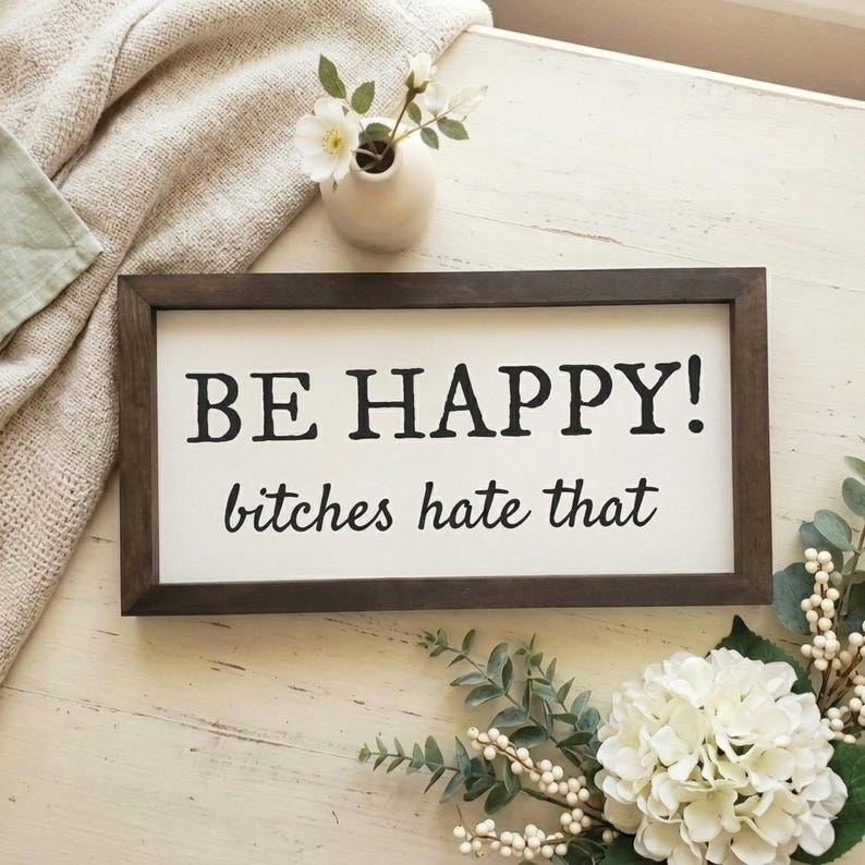 May include: A framed wooden sign with the text "BE HAPPY! bitches hate that" in black letters on a white background. The sign is surrounded by decorative flowers and greenery, with a small white vase holding flowers.