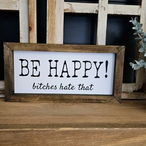 May include: A rectangular wooden framed sign with the words "BE HAPPY!" in bold black letters above the phrase "bitches hate that" in a smaller, cursive font. The sign is on a wooden surface.