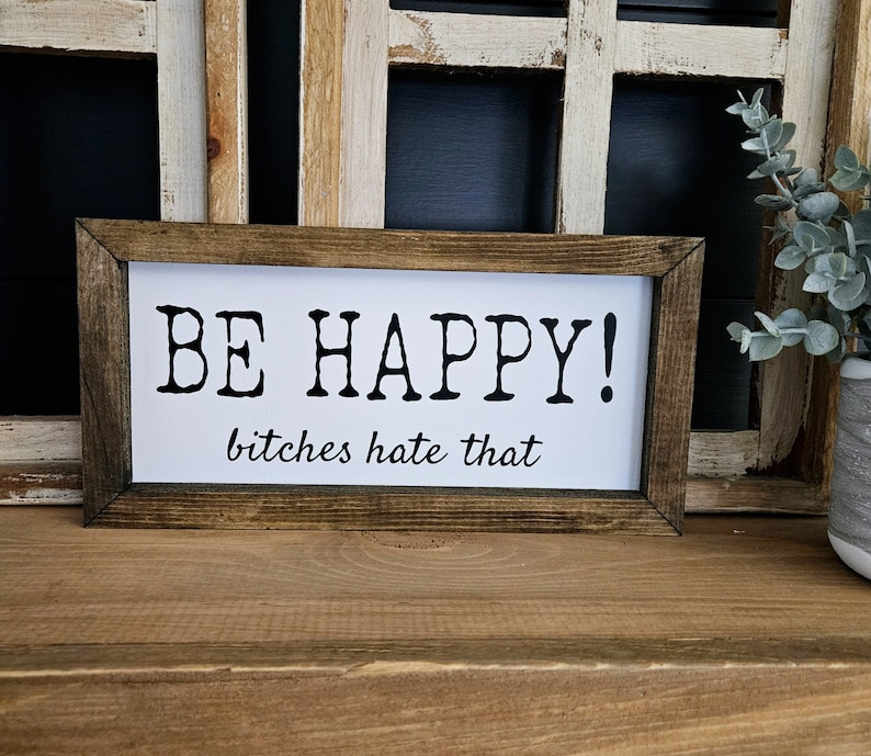 May include: A rectangular wooden framed sign with the words "BE HAPPY!" in bold black letters above the phrase "bitches hate that" in a smaller, cursive font. The sign is on a wooden surface.