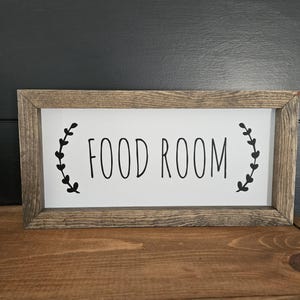 K&ouml;nnte beinhalten: Rechteckiges Holzschild mit den schwarzen Worten "FOOD ROOM" auf wei&szlig;em Hintergrund. Das Schild ist in einem rustikalen, hellbraunen Holz gerahmt. Schwarze Blattmotive flankieren den Text.