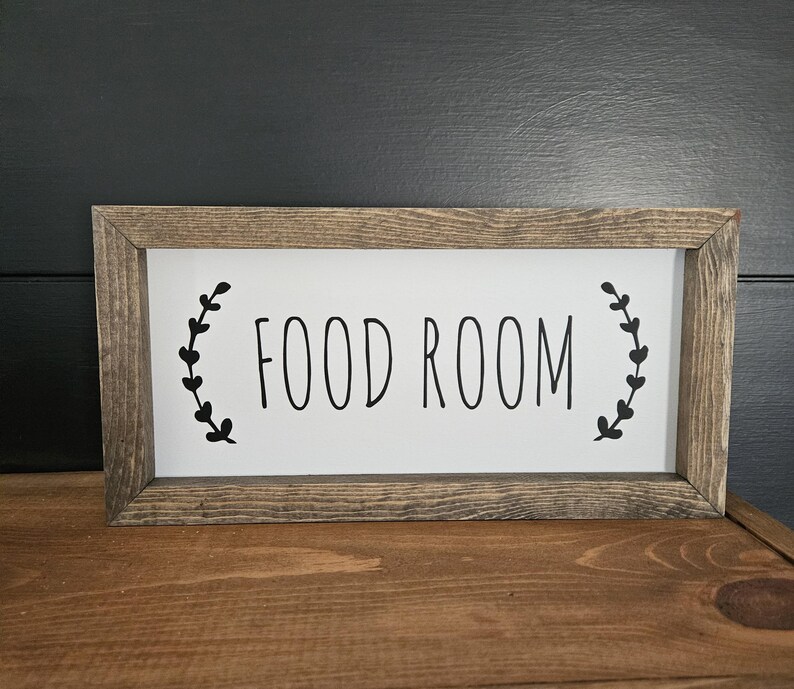 K&ouml;nnte beinhalten: Rechteckiges Holzschild mit den schwarzen Worten "FOOD ROOM" auf wei&szlig;em Hintergrund. Das Schild ist in einem rustikalen, hellbraunen Holz gerahmt. Schwarze Blattmotive flankieren den Text.
