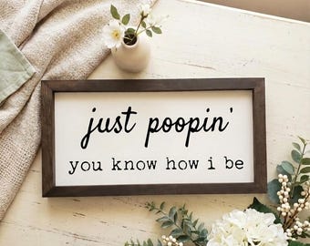 May include: A rectangular wooden sign with a dark brown frame and a white background. The sign features the phrase "just poopin' you know how i be" in black cursive and block letters. The sign is decorated with flowers and a small vase.