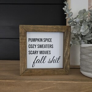 May include: A square wooden sign with a dark brown frame and a white center panel. The text printed in black reads: PUMPKIN SPICE, COZY SWEATERS, SCARY MOVIES, and fall shit in a mix of sans-serif and cursive script fonts.
