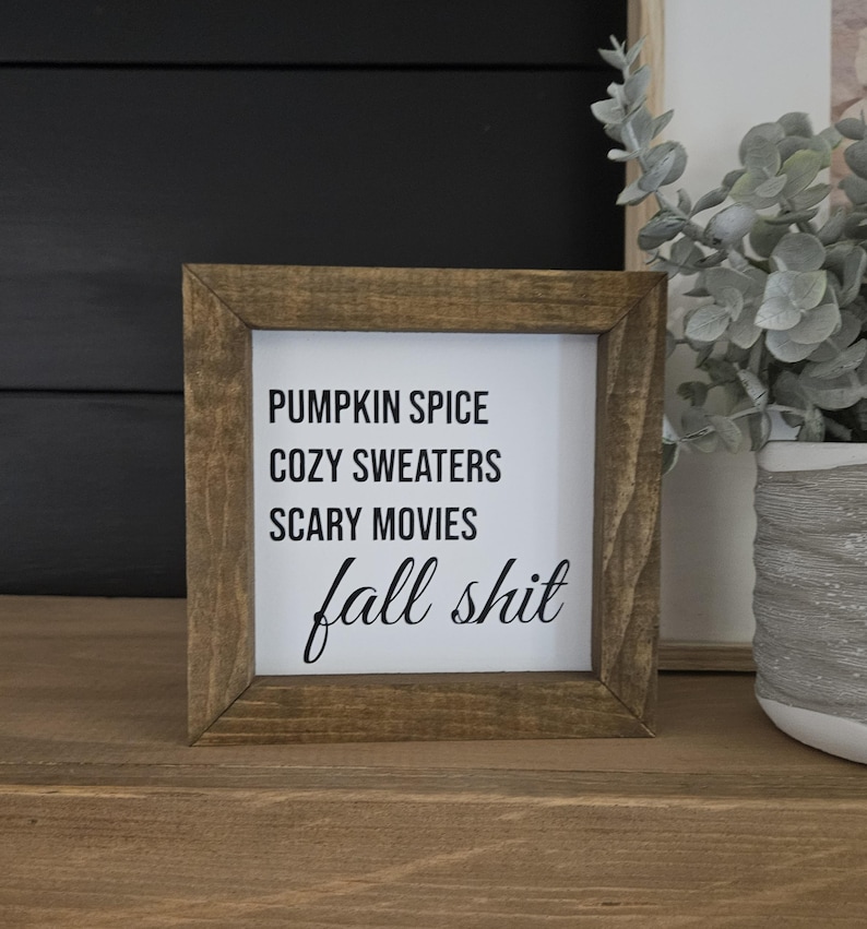 May include: A square wooden sign with a dark brown frame and a white center panel. The text printed in black reads: PUMPKIN SPICE, COZY SWEATERS, SCARY MOVIES, and fall shit in a mix of sans-serif and cursive script fonts.