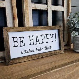 May include: A rectangular wooden sign with a white background and black text that reads "BE HAPPY! bitches hate that." The sign is framed in a dark brown wood and sits on a wooden surface.