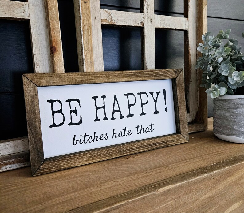 May include: A rectangular wooden sign with a white background and black text that reads "BE HAPPY! bitches hate that." The sign is framed in a dark brown wood and sits on a wooden surface.