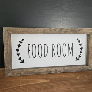K&ouml;nnte beinhalten: Rechteckiges Holzschild mit den schwarzen Worten "FOOD ROOM" auf wei&szlig;em Hintergrund. Das Schild ist in rustikalem, hellbraunem Holz gerahmt und hat schwarze Bl&auml;tter auf beiden Seiten des Textes.