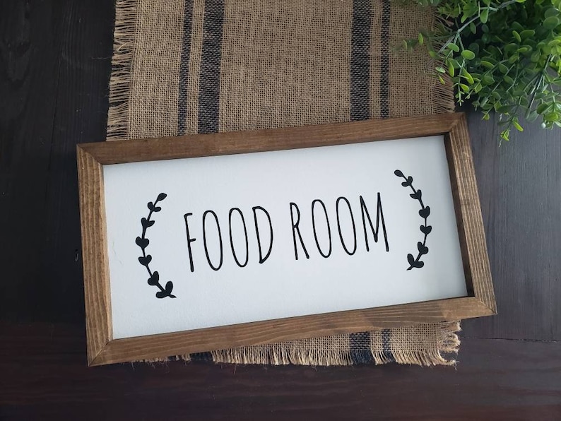 K&ouml;nnte beinhalten: Ein rechteckiges Holzschild mit den schwarzen Worten "FOOD ROOM" auf wei&szlig;em Hintergrund. Das Schild ist mit braunem Holz umrahmt und mit schwarzen Bl&auml;ttern verziert. Das Schild liegt auf einem Jutegewebe.