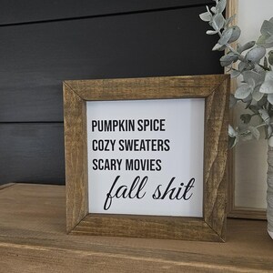 May include: A square wooden sign with a dark brown frame and a white center panel. The text in black reads: PUMPKIN SPICE, COZY SWEATERS, SCARY MOVIES, and fall shit in a cursive script. The sign has a rustic appearance with visible wood grain.