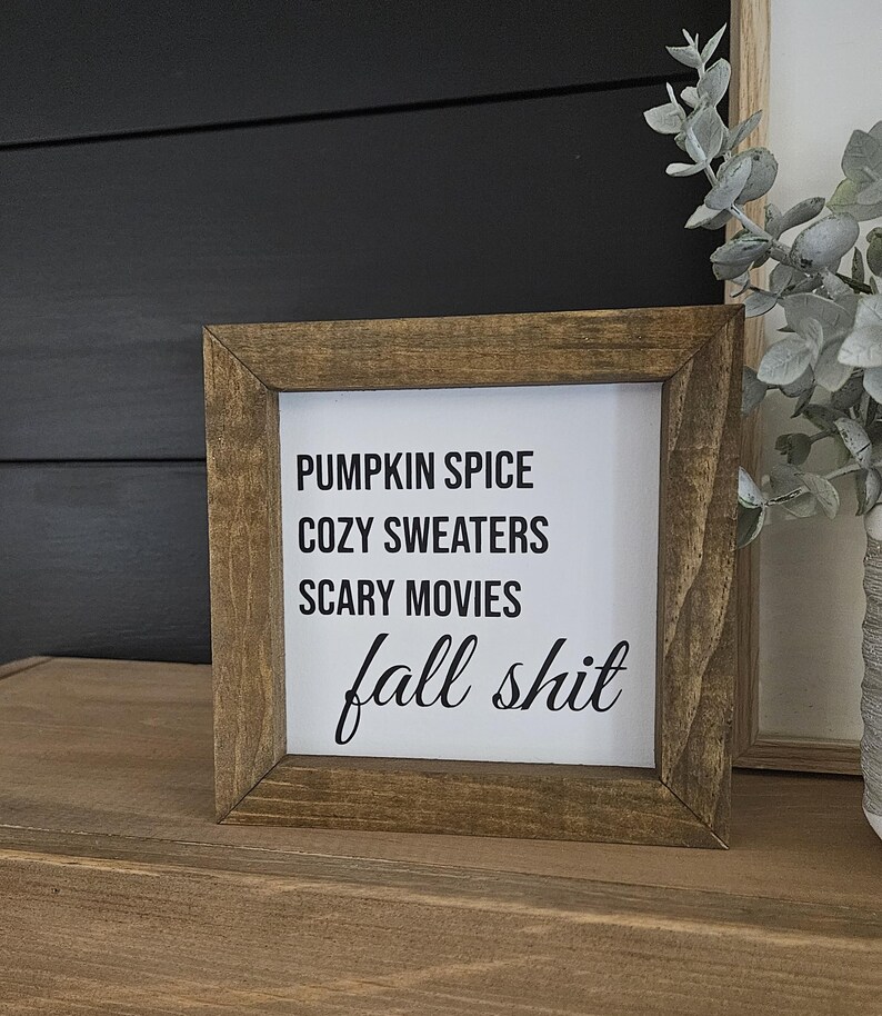 May include: A square wooden sign with a dark brown frame and a white center panel. The text in black reads: PUMPKIN SPICE, COZY SWEATERS, SCARY MOVIES, and fall shit in a cursive script. The sign has a rustic appearance with visible wood grain.