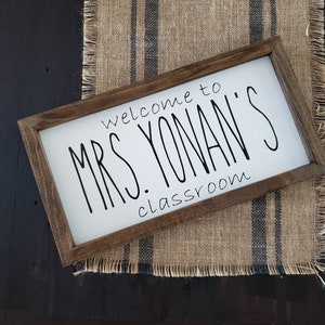 以下が含まれることがあります： 「welcome to MRS. YONAN'S classroom」と黒い手書きフォントで書かれた長方形の木製サイン。サインは、濃い青色のストライプが入ったジュートランナーの上に置かれ、濃い木材の表面に置かれています。