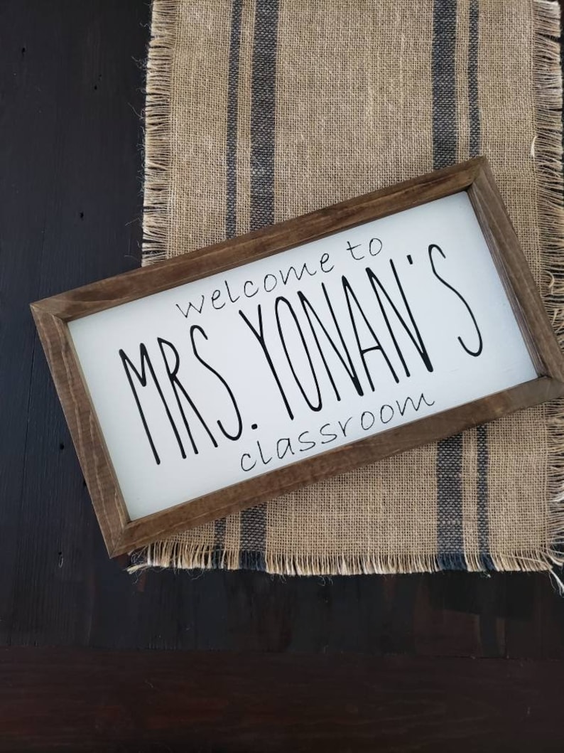 以下が含まれることがあります： 「welcome to MRS. YONAN'S classroom」と黒い手書きフォントで書かれた長方形の木製サイン。サインは、濃い青色のストライプが入ったジュートランナーの上に置かれ、濃い木材の表面に置かれています。