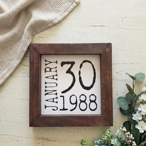 May include: A square wooden framed sign with the text "JANUARY 30 1988" in black lettering on a white background. The frame is a dark brown color. The sign is surrounded by a beige cloth and greenery.