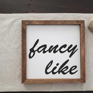 May include: Square wooden framed sign with the words "fancy like" in black cursive script on a white background. The frame is a medium brown color. The sign is resting on a light-colored fabric.