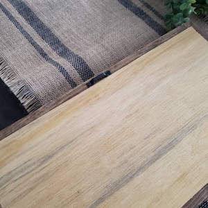 May include: A wooden framed sign with a natural wood grain finish. The frame is a dark brown colour. A metal sawtooth hanger is attached to the back. The sign is resting on a burlap runner with blue stripes.