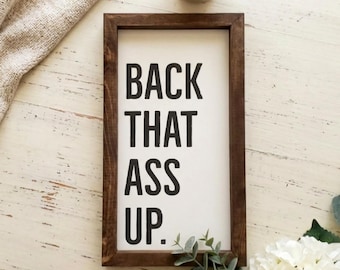 May include: A rectangular wooden sign with a dark brown frame and a white background. The sign features black text in a bold, sans-serif font that reads BACK THAT ASS UP. The sign is oriented vertically and has a clean, simple design.