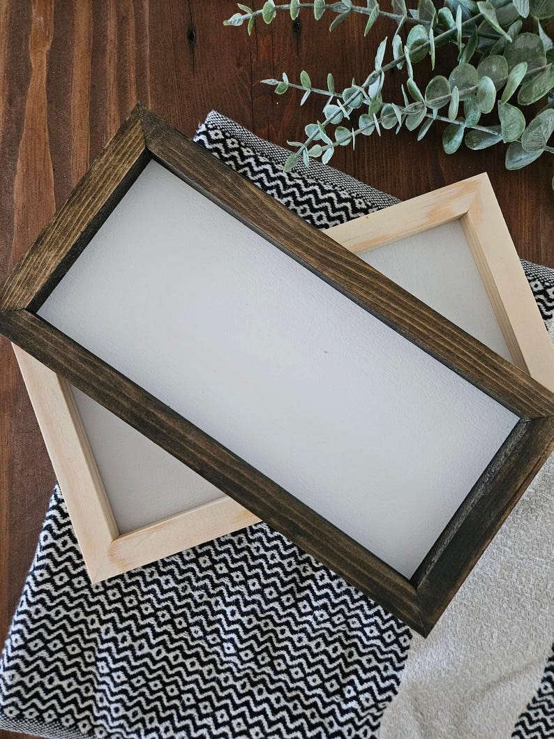 May include: Two rectangular wooden frames with white interiors. One frame has a dark brown stained finish, and the other is a natural light wood color. They are placed on a black and white patterned fabric and a white towel.