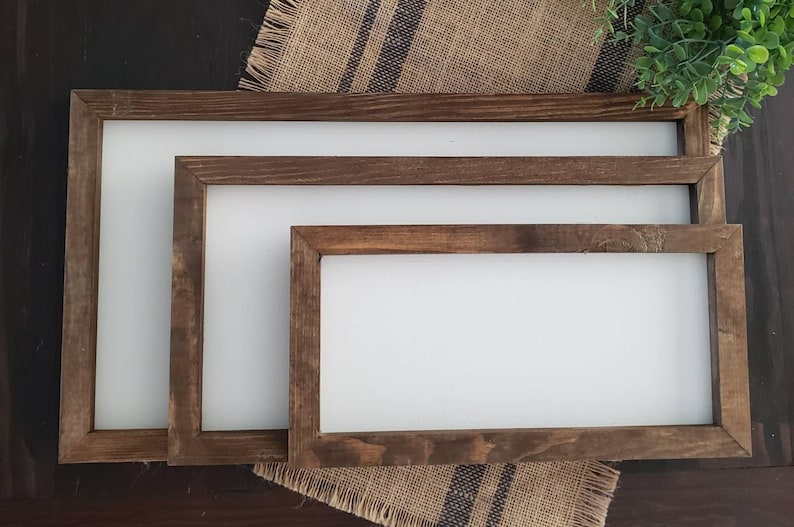K&ouml;nnte beinhalten: Drei rechteckige Holzschilder mit wei&szlig;en Zentren. Die Rahmen sind dunkelbraun. Die Schilder sind ineinander verschachtelt angeordnet, wobei das gr&ouml;&szlig;te Schild hinten und das kleinste vorne ist. Die Schilder befinden sich auf einer Jutefl&auml;che.