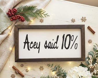 Può includere: Un cartello incorniciato con la scritta "Acey said 10%" in nero. Il cartello è circondato da elementi decorativi tra cui verde, bacche, fiocchi di neve e un bastoncino di cannella, creando un'estetica festosa.