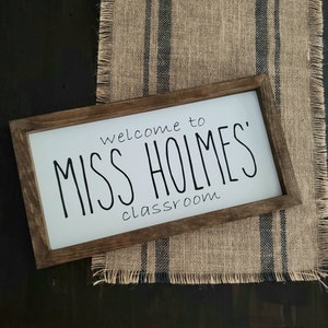 以下が含まれることがあります： 黒い文字で「welcome to MISS HOLMES' classroom」と書かれた長方形の木製サイン。サインは白い背景と濃い茶色のフレームです。青いストライプのジュートランナーの上に置かれています。