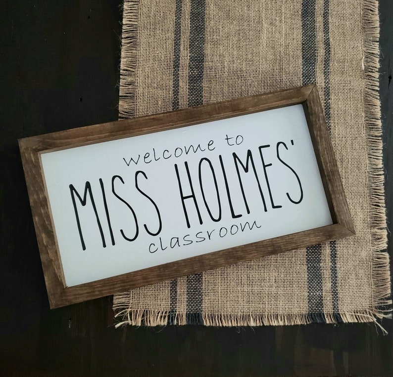 以下が含まれることがあります： 黒い文字で「welcome to MISS HOLMES' classroom」と書かれた長方形の木製サイン。サインは白い背景と濃い茶色のフレームです。青いストライプのジュートランナーの上に置かれています。