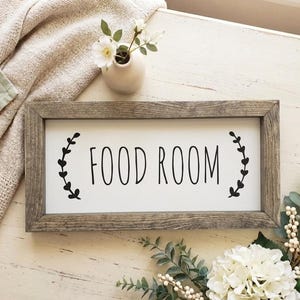 K&ouml;nnte beinhalten: Rechteckiges Holzschild mit den schwarzen Worten "FOOD ROOM" auf wei&szlig;em Hintergrund. Schwarze blattartige Designs flankieren den Text. Das Schild liegt auf einer hellen Holzoberfl&auml;che, mit einer Vase mit Blumen und Gr&uuml;n.