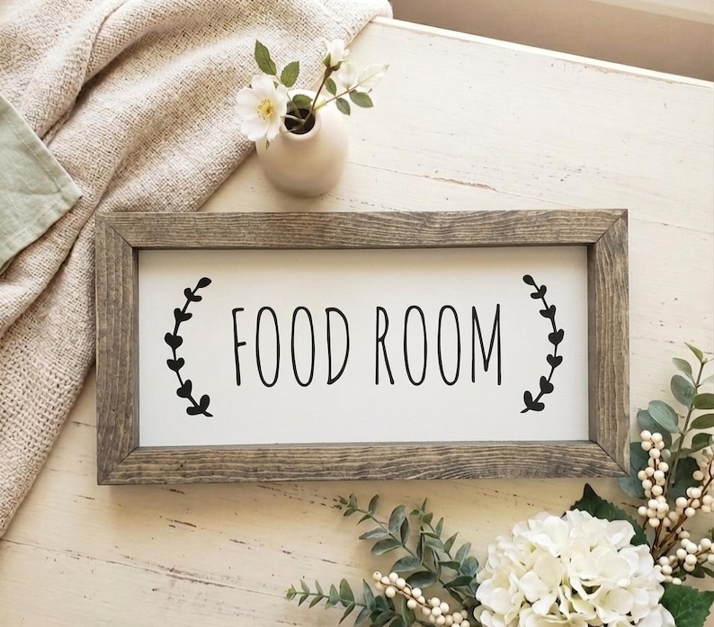 K&ouml;nnte beinhalten: Rechteckiges Holzschild mit den schwarzen Worten "FOOD ROOM" auf wei&szlig;em Hintergrund. Schwarze blattartige Designs flankieren den Text. Das Schild liegt auf einer hellen Holzoberfl&auml;che, mit einer Vase mit Blumen und Gr&uuml;n.