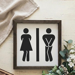 May include: A framed wooden sign with black and white restroom symbols. The sign features a stick figure of a person in a dress and a stick figure of a person in pants. The frame is dark brown, and the background is white.