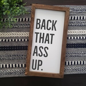 May include: A rectangular wooden framed sign with the phrase "BACK THAT ASS UP" in bold black letters on a white background. The frame is a dark brown colour. The sign is placed on a black and white patterned surface.