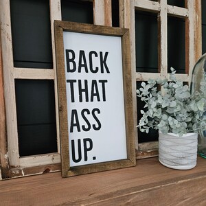 May include: A framed sign with the text "BACK THAT ASS UP" in black on a white background. The sign is in a brown wooden frame, leaning against a distressed wooden window frame.