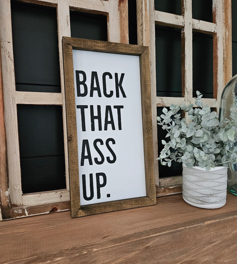 May include: A framed sign with the text "BACK THAT ASS UP" in black on a white background. The sign is in a brown wooden frame, leaning against a distressed wooden window frame.