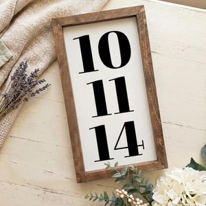 May include: A rectangular wooden framed sign with the numbers 10, 11, and 14 in bold black font on a white background. The frame is a dark brown color. The sign is decorated with sprigs of lavender and white flowers.