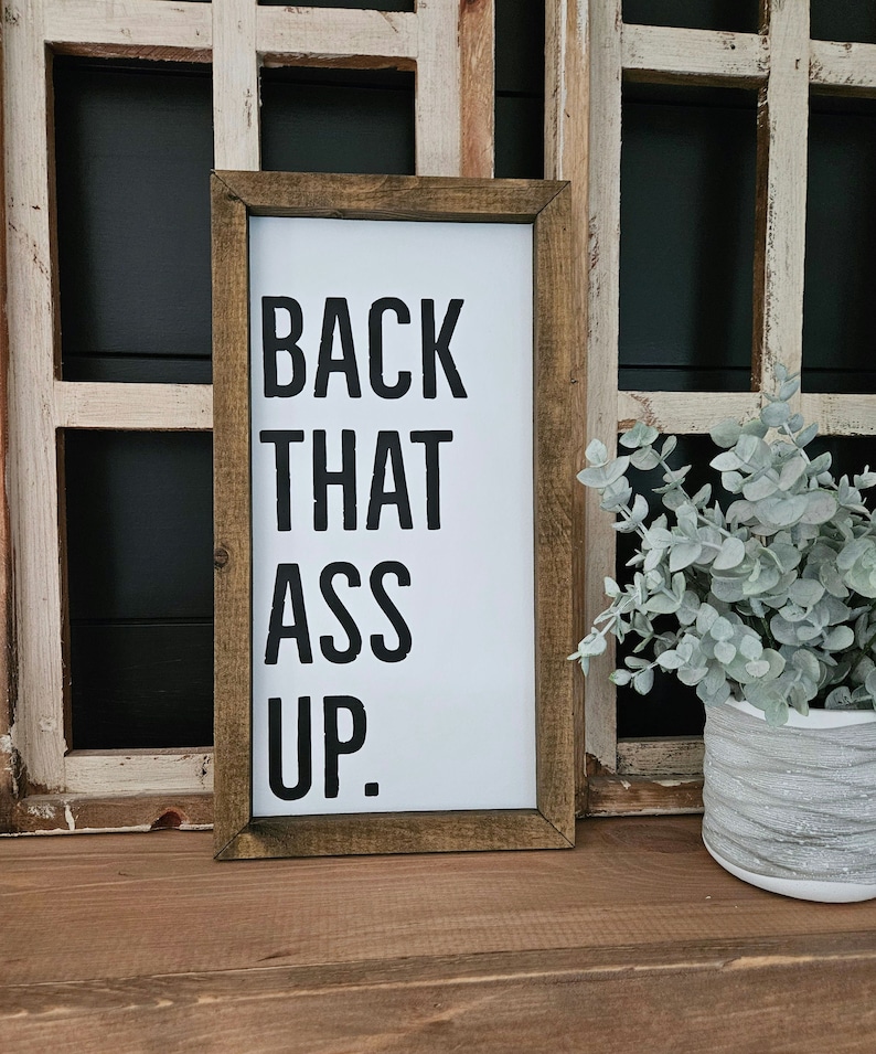 May include: A rectangular wooden framed sign with the text "BACK THAT ASS UP" in bold black letters on a white background. The sign is displayed on a wooden surface. A decorative plant in a white pot is in the background.