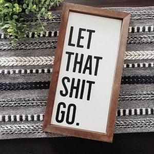 May include: A rectangular wooden framed sign with the text "LET THAT SHIT GO." in bold black letters on a white background. The sign is placed on a black and white patterned runner with a green plant in the background.