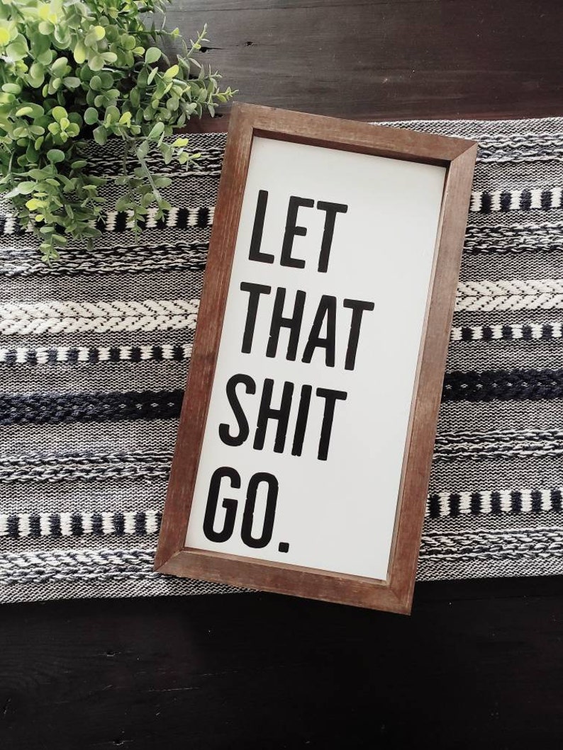 May include: A rectangular wooden framed sign with the text "LET THAT SHIT GO." in bold black letters on a white background. The sign is placed on a black and white patterned runner with a green plant in the background.