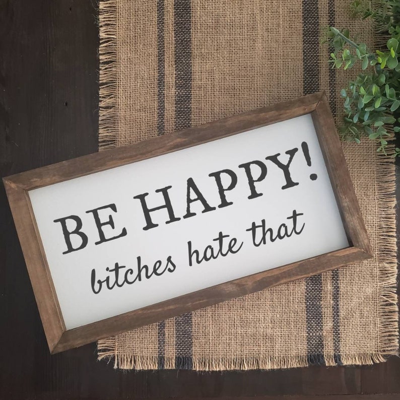 May include: A rectangular wooden sign with the words "BE HAPPY! bitches hate that" in black lettering on a white background. The sign is framed in dark brown wood and rests on a burlap runner.