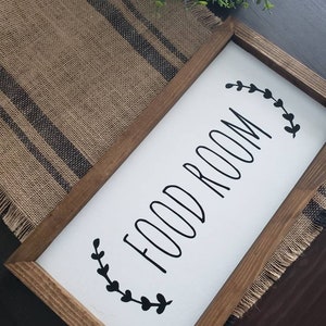 K&ouml;nnte beinhalten: Holzgerahmtes Schild mit den Worten "FOOD ROOM" in schwarzer Schrift auf wei&szlig;em Hintergrund. Das Schild ist mit schwarzen Bl&auml;ttern verziert und hat einen rustikalen, l&auml;ndlichen Stil.