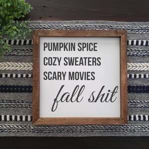 May include: A square wooden sign with a dark brown frame and a white background. The text in black reads: PUMPKIN SPICE, COZY SWEATERS, SCARY MOVIES, and fall shit in a cursive script. The sign is displayed on a dark surface with a woven runner.