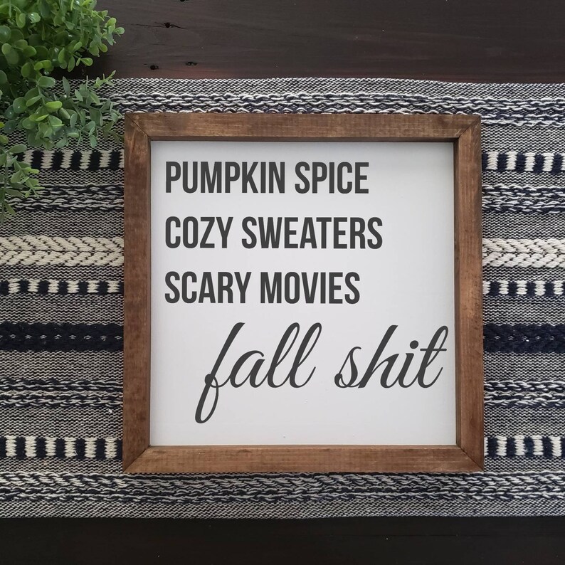 May include: A square wooden sign with a dark brown frame and a white background. The text in black reads: PUMPKIN SPICE, COZY SWEATERS, SCARY MOVIES, and fall shit in a cursive script. The sign is displayed on a dark surface with a woven runner.